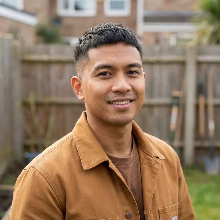 Smiling man with short dark hair wearing a tan jacket in a backyard garden.